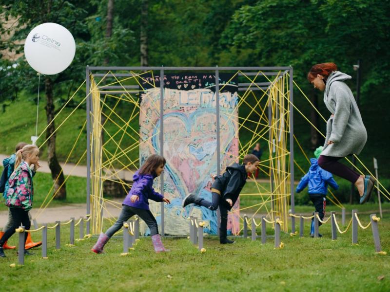 Bērnu grupa un pieaugušais parkā priecīgi skrien un lec krāsainas sirds formas sienas gleznojuma priekšā. Kreisajā pusē redzams balons, un tos ieskauj zaļi koki.