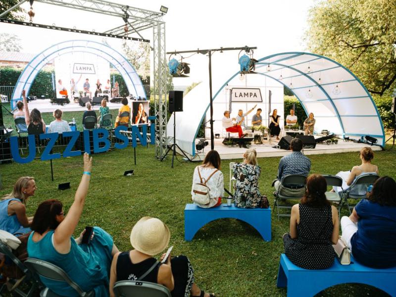 Āra paneļdiskusija festivālā zem izliektas nojumes. Dalībnieki sēž uz soliņiem un komunicē ar runātājiem uz skatuves. Gaiša, dzīvīga atmosfēra.