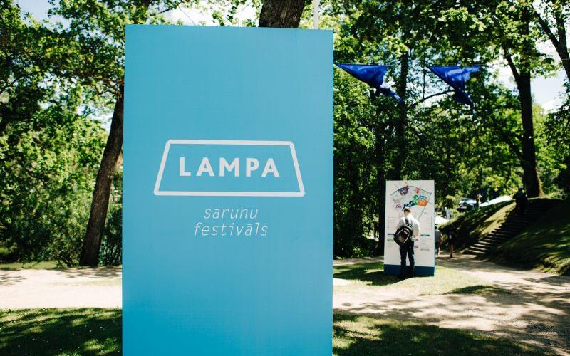 Saulainā parkā stāv liela zila zīme ar uzrakstu "LAMPA sarunu festivāls". Netālu pastaigājas cilvēki, un starp sulīgi zaļiem kokiem plīvo zili karogi, radot svētku atmosfēru.