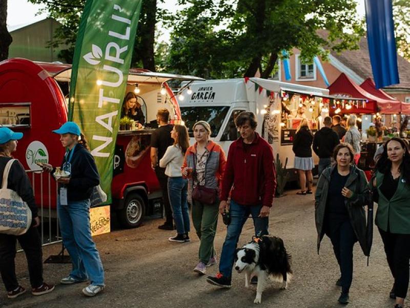 Cilvēki pastaigājas pa festivāla ēdināšanas zonu svētku atmosfērā. Gar taku stāv ēdienu furgoni ar krāsainiem plakātiem un zīmēm.