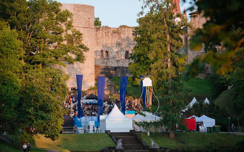 Cēsu pils pilsdrupām priekšā notiek brīvdabas festivāls ar ziliem karogiem, baltām teltīm un dzīvīgu pūli, ko ieskauj sulīga zaļums.