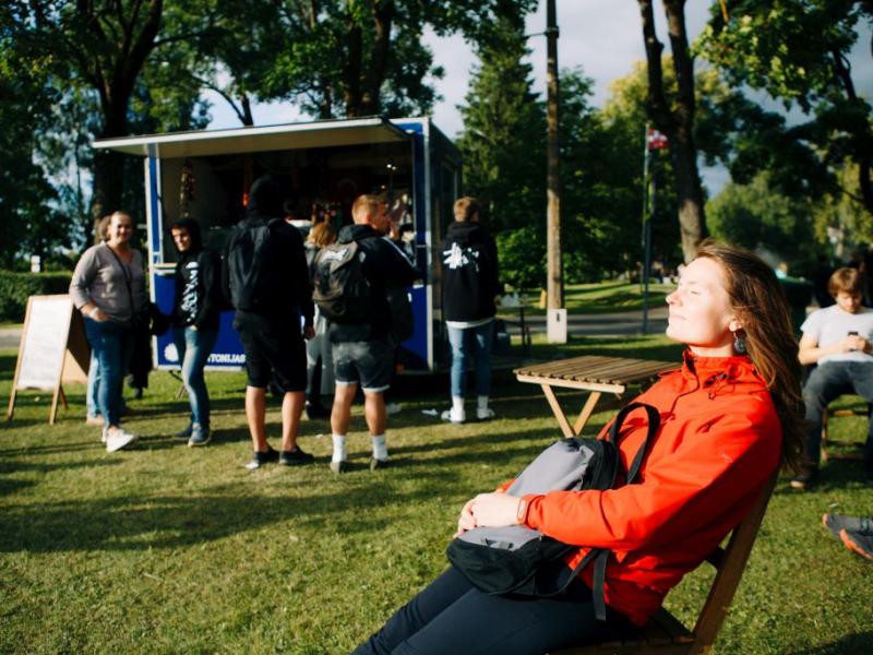 Sieviete sarkanā jakā atpūšas saulainā dienā parkā ar aizvērtām acīm un smaidot. Fonā cilvēki pulcējas pie zila ēdienu furgona starp zaļiem kokiem.