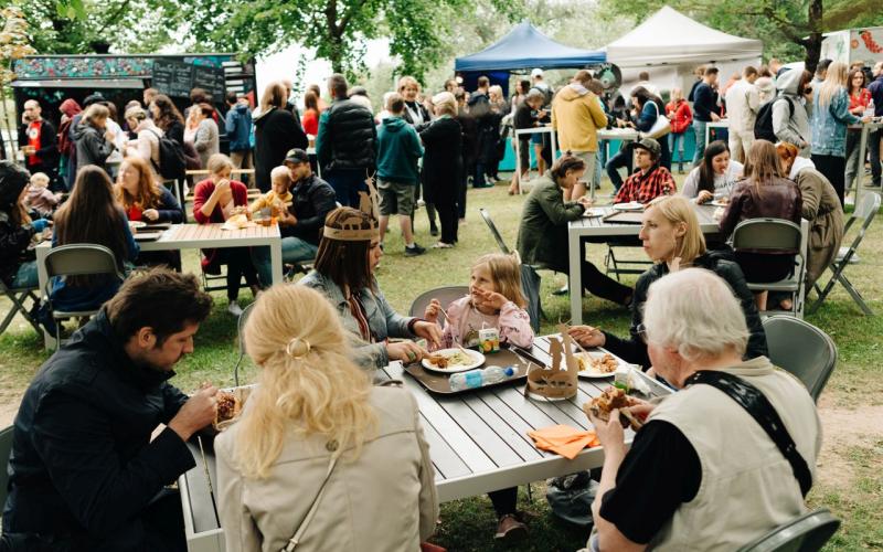 Festivāls, kurā visu vecumu cilvēki sēž pie galdiem un bauda maltītes zem kokiem. Fonā ir ēdienu furgoni un teltis, kas rada dzīvīgu atmosfēru.