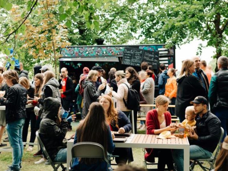 Festivāls, kurā visu vecumu cilvēki sēž pie galdiem un bauda maltītes zem kokiem. Fonā ir ēdienu furgoni un teltis, kas rada dzīvīgu atmosfēru.