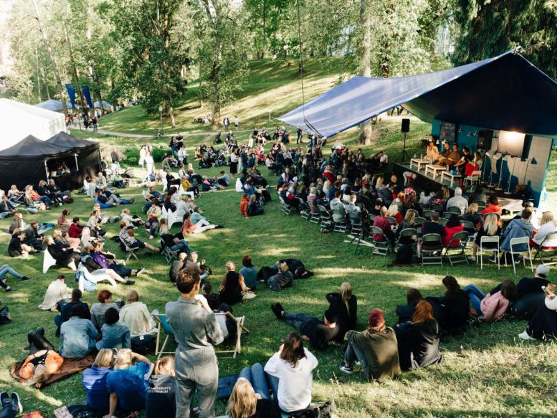Festivāla aina brīvā dabā parkā ar zilu telti, kurā notiek paneļdiskusija. Dažāda auditorija sēdvietās, relaksēta atmosfēra koku ieskautā.