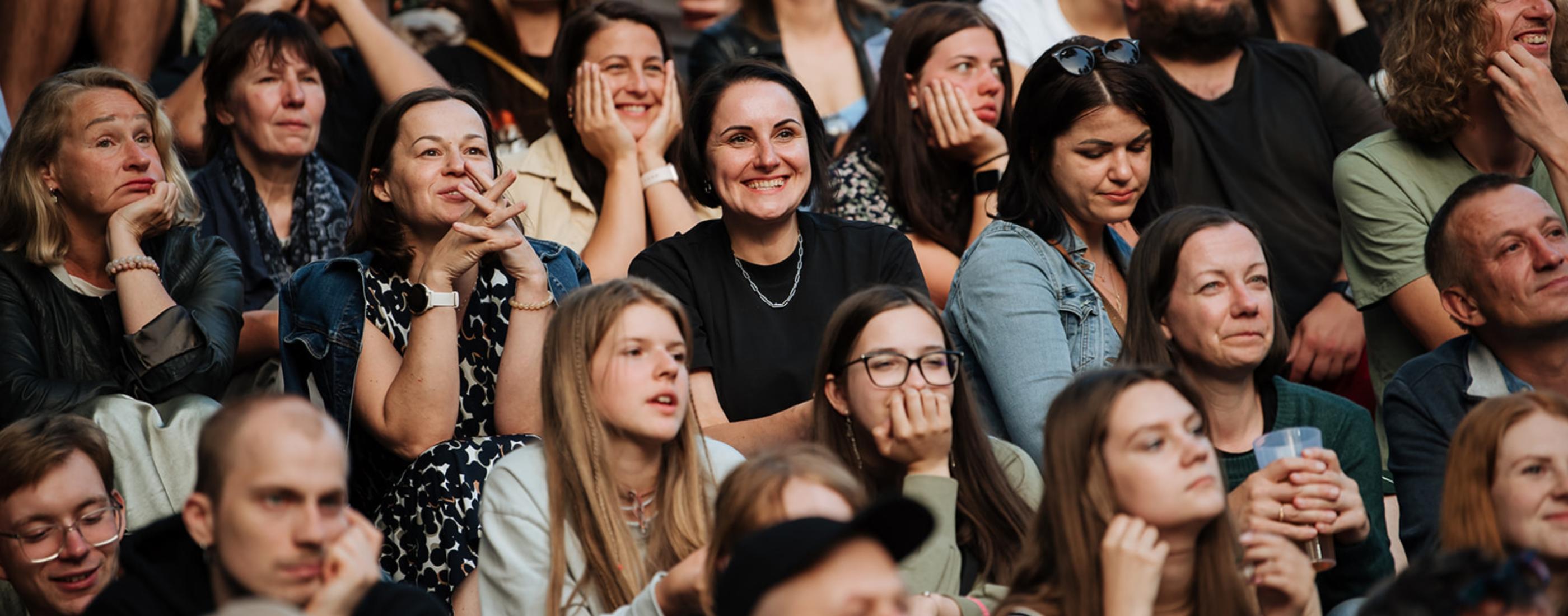 Liels, daudzveidīgs pūlis sēž cieši saspiedies, uzmanīgi vērojot ar ieinteresētām un smaidošām sejām. Atmosfēra ir dzīvīga un aizraujoša.