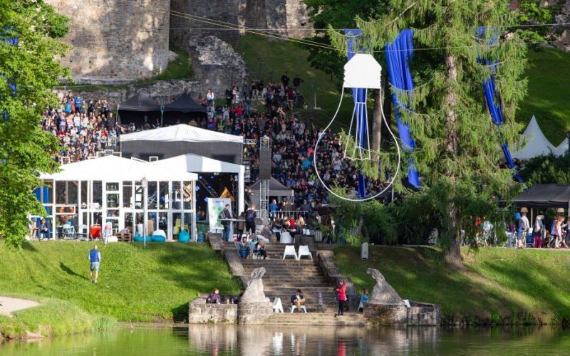 Brīvdabas festivāla aina ar lielu pūli, kas pulcējas ap baltām teltīm un zilām dekorācijām. Liels koks, kāpnes un mierīgs ezers pastiprina dzīvīgo atmosfēru.