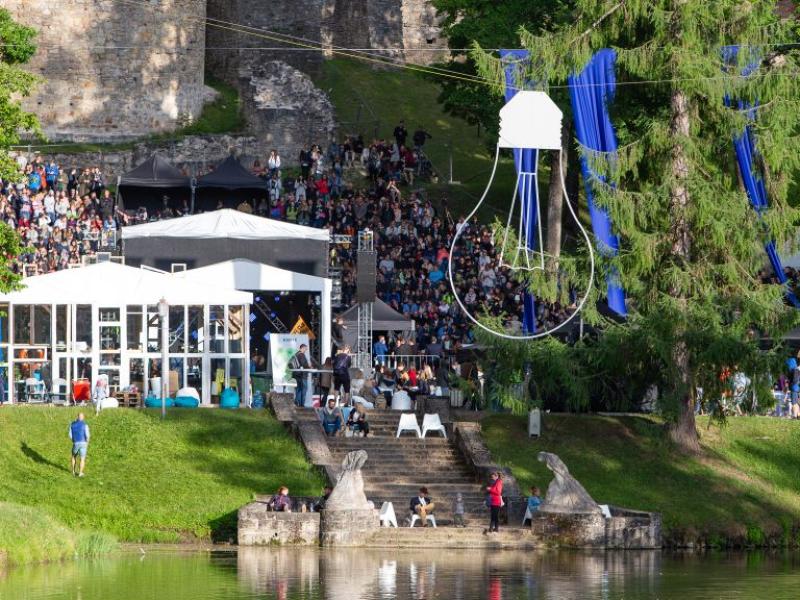 Brīvdabas festivāla aina ar lielu pūli, kas pulcējas ap baltām teltīm un zilām dekorācijām. Liels koks, kāpnes un mierīgs ezers pastiprina dzīvīgo atmosfēru.
